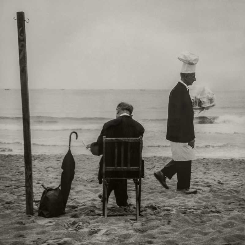 Fotografía de un caballero sentado y un camarero caminando junto al mar